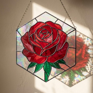 May include: A stained glass hexagon with a red rose design. The rose has detailed petals and green leaves. The glass has a shimmering, iridescent quality. A metal chain is attached for hanging.