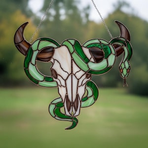 May include: Stained glass art piece featuring a bull skull with brown horns and a green snake wrapped around it. The skull is white and brown, and the snake has green and brown segments. The piece is hanging from a silver chain.