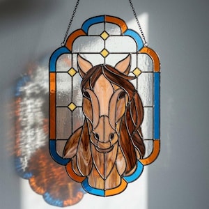 May include: A stained glass suncatcher featuring a horse head portrait in shades of brown and tan. The piece is framed by a decorative border of blue and orange glass segments with small yellow diamond accents, all held together by dark metal solder.