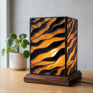Könnte beinhalten: Eine rechteckige Tischlampe aus Buntglas mit einem schwarz-orangen Tigerstreifenmuster. Die Lampe ist mit dunklen Metalllötstellen zwischen den Glasscheiben konstruiert und auf einem quadratischen, dunkel gehaltenen Holzsockel montiert.