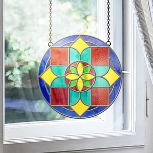May include: A circular stained glass window suspended by a chain. The design incorporates a central floral motif and geometric shapes in red, yellow, green, and blue. The window is framed in dark grey metal.