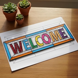 Könnte beinhalten: Ein farbenfrohes Schild im Glasmalerei-Stil mit dem Wort "WELCOME" in fetten, mehrfarbigen Buchstaben. Das Schild befindet sich auf weißem Papier mit einem schwarzen Umrissmuster und ist von orangefarbenen, blauen und violetten Rändern umgeben. Im Hintergrund sind drei kleine Sukkulenten in Töpfen.