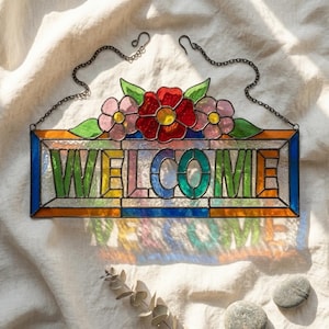 May include: A rectangular stained glass sign featuring the word WELCOME in multicolored block letters. The piece is topped with three glass flowers in red and pink with green leaves and includes a black metal chain for hanging purposes.