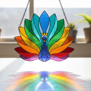 May include: A stained glass suncatcher shaped like a peacock with a fanned tail. The piece features vibrant segments of red, orange, yellow, green, teal, blue, and purple glass held together by dark metal lines, suspended by a thin metal chain.