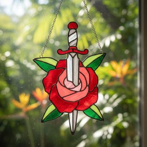 May include: Stained glass art piece featuring a red rose pierced by a silver sword. The rose has pink and red petals with green leaves. The piece is suspended by a silver chain, creating a decorative window hanging.