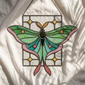 May include: A stained glass suncatcher featuring a green and pink moth centred within a rectangular frame. The moth has dark green, light green, and teal wings with orange accents. The background consists of clear textured glass and yellow stars.