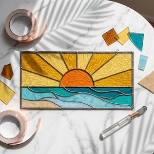 Könnte beinhalten: Ein Glasfenster mit einem Sonnenuntergang über dem Meer. Die Sonne ist orange, mit gelben Strahlen. Das Meer ist blau und türkis. Kupferband und Glasscherben umgeben das Fenster. Ein Glasschneider und Rollen mit Kupferband sind ebenfalls vorhanden.