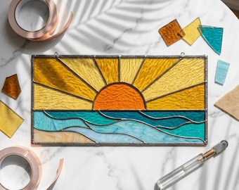 Op de afbeelding: Een glas-in-loodpaneel met een zonsondergang boven de oceaan. De zon is oranje, met gele stralen. De oceaan is blauw en turquoise. Koperband en glasstukken omringen het paneel. Een glassnijder en rollen koperband zijn ook aanwezig.