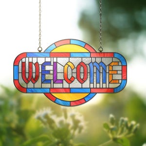 May include: A stained glass "WELCOME" sign with a colourful design. The sign features a rectangular shape with rounded edges, a red border, and a blue outline. The word "WELCOME" is written in bold, multi-coloured letters. The sign hangs from two silver chains.