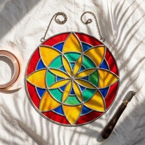 May include: A vibrant stained glass window panel with a floral pattern. The circular design incorporates red, yellow, blue, and green glass, set within a silver metal frame and suspended by chains. Copper tape and a glass cutter are also present.
