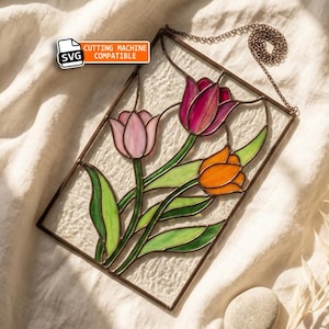 Puede incluir: Un panel rectangular de vidrieras con tres tulipanes en tonos rosa claro, magenta intenso y naranja con hojas verdes. La pieza está enmarcada en metal, incluye una cadena para colgar y muestra una etiqueta que dice SVG CUTTING MACHINE COMPATIBLE.