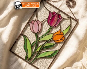May include: A rectangular stained glass panel featuring three tulips in shades of light pink, deep magenta, and orange with green leaves. The piece is framed in metal, includes a hanging chain, and displays a label reading SVG CUTTING MACHINE COMPATIBLE.