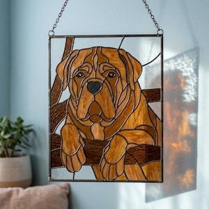 May include: A rectangular stained glass suncatcher featuring a tan and brown dog with a black nose and eyes, resting its paws on a horizontal wooden beam. The piece is framed in dark metal and hangs from a silver chain against a light blue wall.