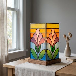 Peut inclure: Une lampe de table rectangulaire en vitrail présentant un motif floral avec des pétales roses, verts et bleus sur un fond de ciel jaune et orange. La lampe est construite avec des lignes de plomb en métal sombre et repose sur une base en bois.