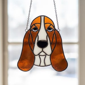 May include: Stained glass basset hound dog suncatcher with brown, white, and black details. The dog's face is framed by a black border and hangs from a metal chain. A decorative piece for dog lovers.
