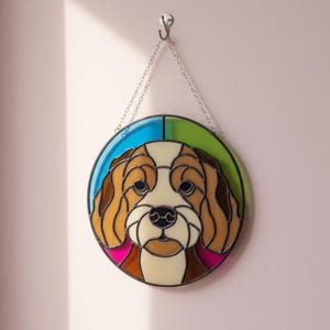 May include: A round stained glass dog portrait with a chain for hanging. The dog has brown and white fur, a black nose, and dark eyes. The background is divided into blue, green, and pink sections.