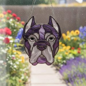 May include: A decorative stained glass dog head in purple, white, and grey, suspended by a chain. The dog's face is crafted with geometric shapes. The background is a blurred garden scene, adding a soft contrast to the artwork.