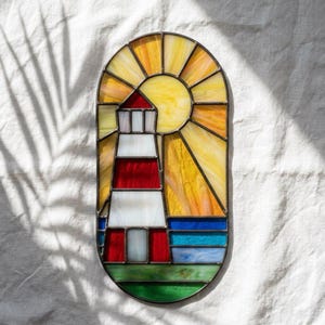May include: An oval stained glass panel featuring a lighthouse with red and white horizontal stripes. The background depicts a yellow sun with radiating beams, a blue sea, and a green foreground, all held together by dark metal lead lines.