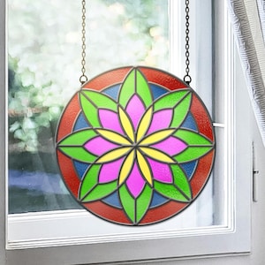 May include: A circular stained glass window, suspended by a chain. It showcases a floral pattern with a red border, blue and green petals, and a pink and yellow central design. The window is positioned in front of a window.
