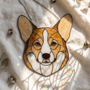 Op de afbeelding: Gebrandschilderd glazen corgi-hoofd in bruin, wit en beige. Het gezicht van de hond heeft bruine ogen en een donkerbruine neus. Een zilveren ketting is bevestigd om op te hangen. Een decoratief kunstwerk.
