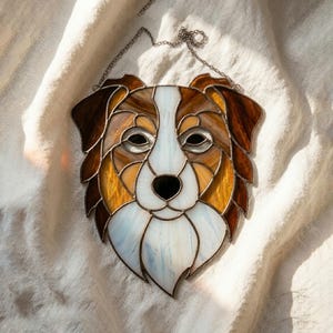May include: Stained glass dog face with brown, tan, and white sections. The dog's face is made of multiple pieces of glass, with a chain for hanging. The dog's eyes and nose are black. The glass has a textured appearance.