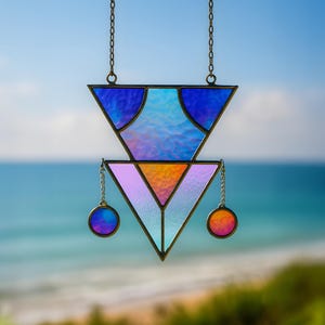 May include: A geometric stained glass suncatcher. The design features two triangles, one above the other, in shades of blue, purple, orange, and pink. Two small, round accents hang from the bottom triangle. The suncatcher is suspended by chains.
