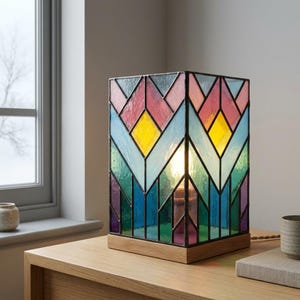 Könnte beinhalten: Eine rechteckige Tischleuchte aus Buntglas mit einem geometrischen Zickzackmuster in leuchtendem Gelb, Rosa, Blau und Grün. Die Glasscheiben sind mit dunklen Metallnähten verbunden und die Leuchte steht auf einem hellen Holzsockel.