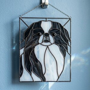 Puede incluir: Un panel rectangular de vidriera que presenta un retrato estilizado de un perro Chin japonés. La pieza está elaborada con segmentos de vidrio blanco y negro unidos por líneas de soldadura de metal oscuro y cuelga de una fina cadena de metal plateado.