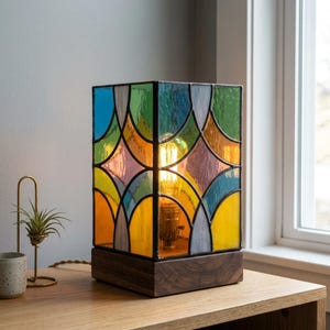 Puede incluir: Una lámpara de mesa rectangular de vidrieras con un patrón geométrico de paneles de vidrio azul, verde, amarillo y morado unidos por perfiles metálicos oscuros. La lámpara se asienta sobre una base de madera oscura y contiene una bombilla.
