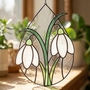 May include: A stained glass panel in a teardrop shape, depicting two white snowdrop flowers with green stems and accents. The glass is clear and textured, with black framing. A decorative piece.