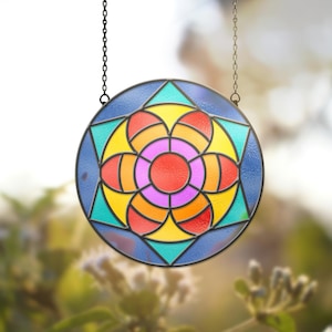 May include: A circular stained glass window with a floral design. The window features a central red circle surrounded by a flower pattern in shades of pink, orange, yellow, and teal. It is framed by a blue border and hangs from a black chain.