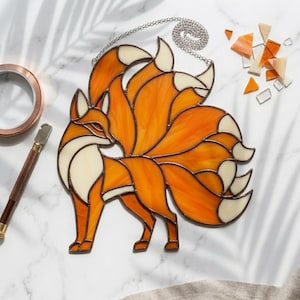 May include: Stained glass fox with nine tails, featuring orange and cream-colored glass. The fox is depicted in a standing pose, with a silver chain for hanging. The artwork is likely for home decor.