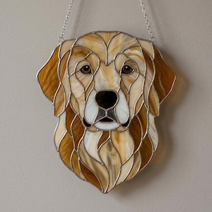May include: Stained glass depiction of a Golden Retriever dog's head. The artwork features shades of brown, tan, and white glass pieces, outlined with silver solder. The dog's eyes are brown, and a silver chain is attached for hanging.