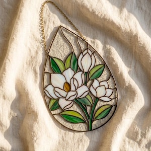 Puede incluir: Vidriera con flores de magnolia en forma de lágrima. La obra presenta flores de magnolia blancas con centros amarillos y hojas verdes, enmarcadas por un contorno de metal oscuro. Una cadena dorada está adjunta para colgar.
