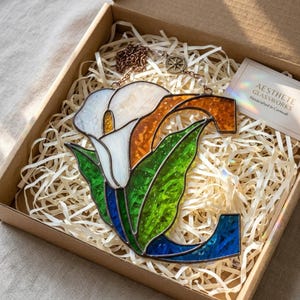 May include: A stained glass letter C featuring a white calla lily with a yellow center, green leaves, and orange and blue glass accents. The piece includes a copper-colored metal chain for hanging and is presented in a cardboard gift box.