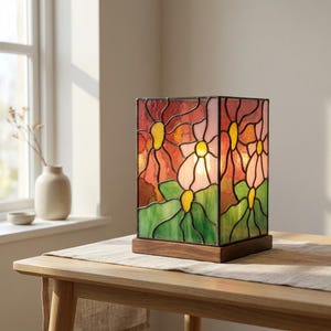 May include: A square stained glass table lamp featuring a floral pattern with red, pink, and yellow petals against green leaves. The lamp is constructed with dark metal soldering lines and is mounted on a rectangular wooden base, emitting a warm glow.