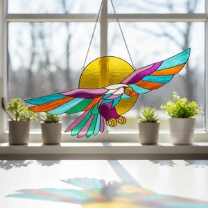 May include: A stained glass eagle in flight, with a golden sun in the background. The eagle features vibrant colors including teal, orange, purple, and pink. It hangs from a chain, perfect for a window decoration.