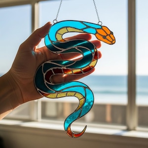 Op de afbeelding: Een gebrandschilderde slangen-zonnevanger in tinten blauw, blauwgroen, oranje, geel en rood. De slang wordt opgehangen aan een ketting. De zonnevanger wordt voor een raam met uitzicht op de oceaan gehouden.
