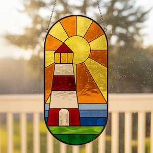 Puede incluir: Una vidriera que representa un faro. El faro es rojo y blanco, con un tejado rojo. El sol es amarillo y naranja, y el mar es azul. La ventana está enmarcada en negro y cuelga de una cadena.