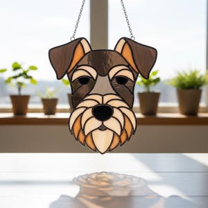 May include: Stained glass Schnauzer dog head hanging decoration. The dog's face features a black nose, brown ears, and a light brown and cream-colored muzzle. The piece is suspended by a chain and is designed to hang in a window.