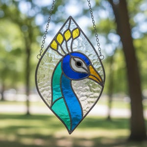 Pode incluir: Enfeite de janela de pavão em vitral. O pavão é retratado em tons de azul, verde, amarelo e branco, com uma coroa amarela. O vidro é colocado em uma moldura em forma de diamante com uma corrente para pendurar.
