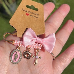 May include: A gold-tone safety pin brooch featuring a light pink satin bow, a braided pink cord, and three dangling charms: an oval religious pendant with a pink center, a pair of baby feet, and a cross with a small pink stone in the center.