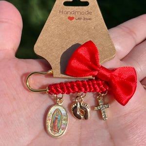 May include: A red bow and charm pin with a gold safety pin clasp. The pin features a red braided cord and three dangling charms: an oval pendant with a religious image, baby footprints, and a cross.