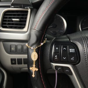 May include: A gold-colored rosary hanging from a black leather steering wheel. The rosary features a cross pendant and an oval pendant with a religious figure. The steering wheel has red stitching.