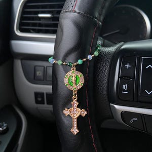 May include: A gold-colored car charm featuring a cross pendant with clear rhinestones and a circular pendant with a green design. The charm is attached to a beaded strand with green and blue beads. The charm hangs from a black steering wheel.