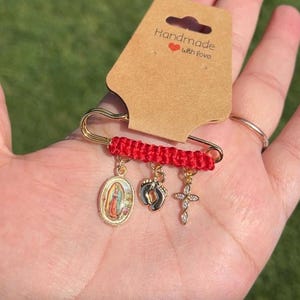 May include: A gold-colored safety pin with a red woven accent and three dangling charms. Charms include an oval religious image, a baby footprint, and a cross with clear stones. The attached tag reads "Handmade with love."
