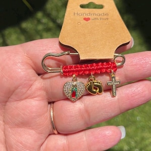 May include: A gold-tone safety pin featuring a red woven fabric band and three dangling charms: a heart-shaped pendant with a central figure and clear stones, a pair of gold baby feet, and a small cross. The pin is attached to a brown card.