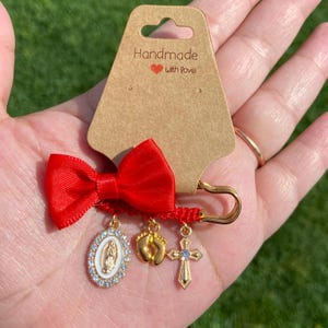 Puede incluir: Un llavero hecho a mano con un lazo rojo y dijes dorados. Los dijes incluyen un colgante de la Virgen María, pies de bebé y una cruz. La etiqueta dice "Handmade with Love."