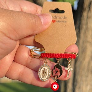 May include: A gold-colored safety pin with a red cord and various charms. Charms include an oval pendant with a religious figure, a mother and child silhouette, a cross, and a red evil eye. The pin is attached to a brown tag that reads "handmade with love."
