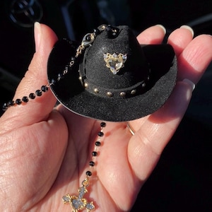 May include: A miniature black cowboy hat with gold studs and a heart-shaped jewel. A black beaded chain is attached to the hat, leading to a gold cross pendant with clear gemstones. The hat is held in a hand.